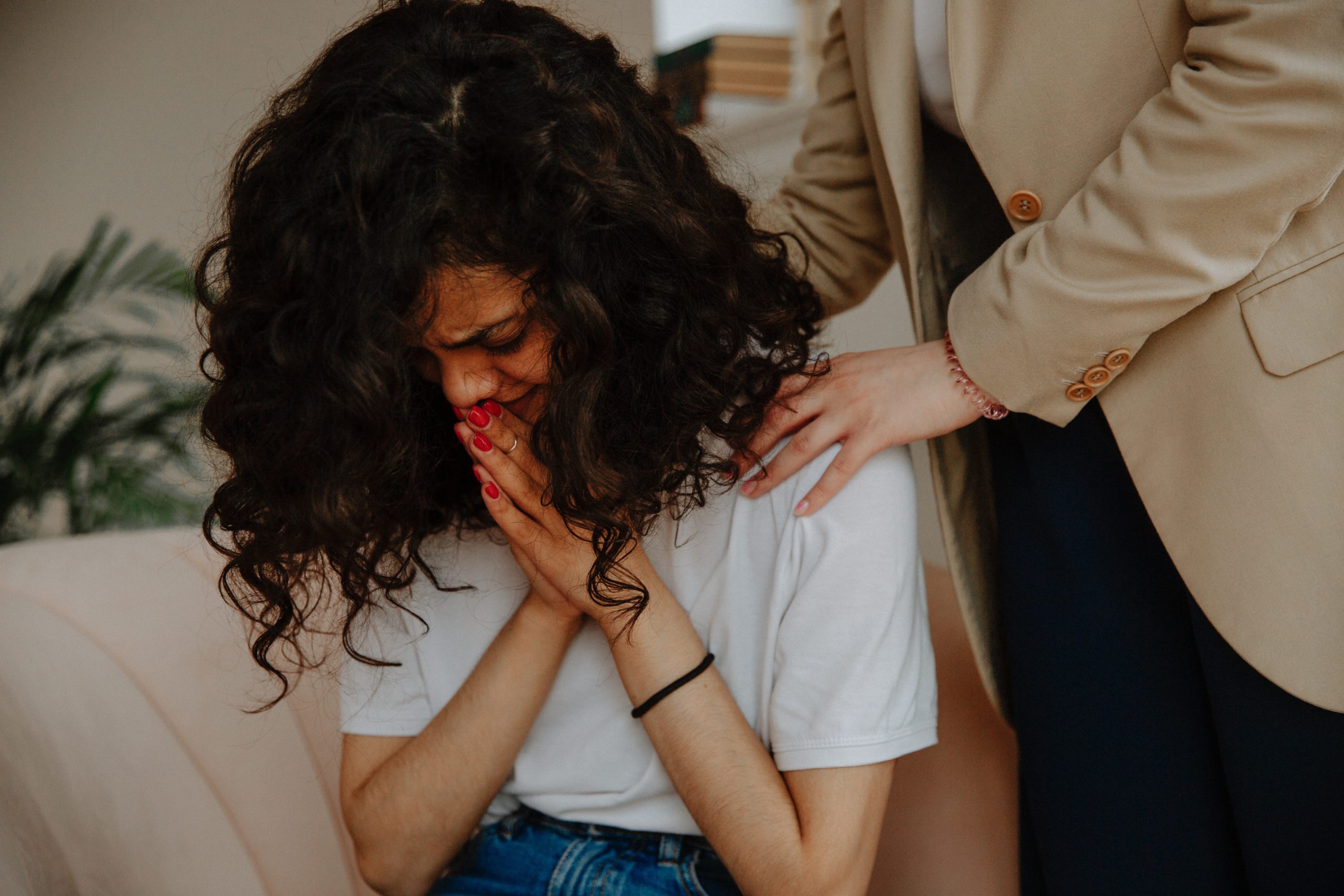 Teen sitting on a couch crying while someone stands over her consoling her