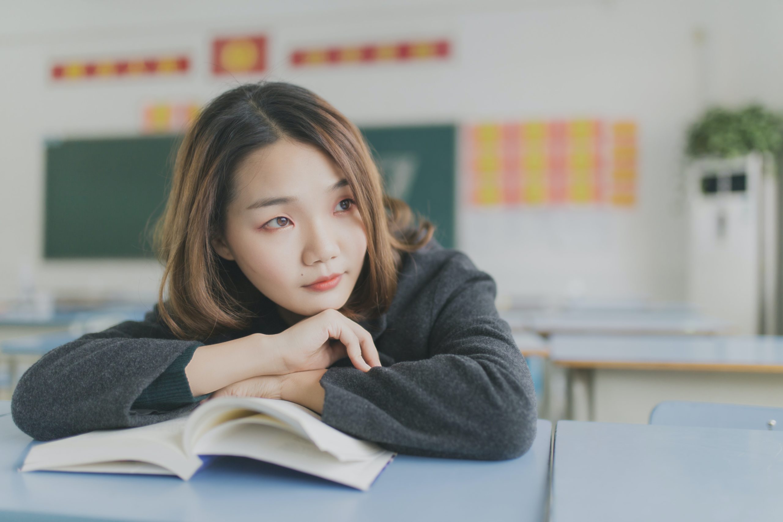 Girl resting her head on a book, looking out the window in a classroom