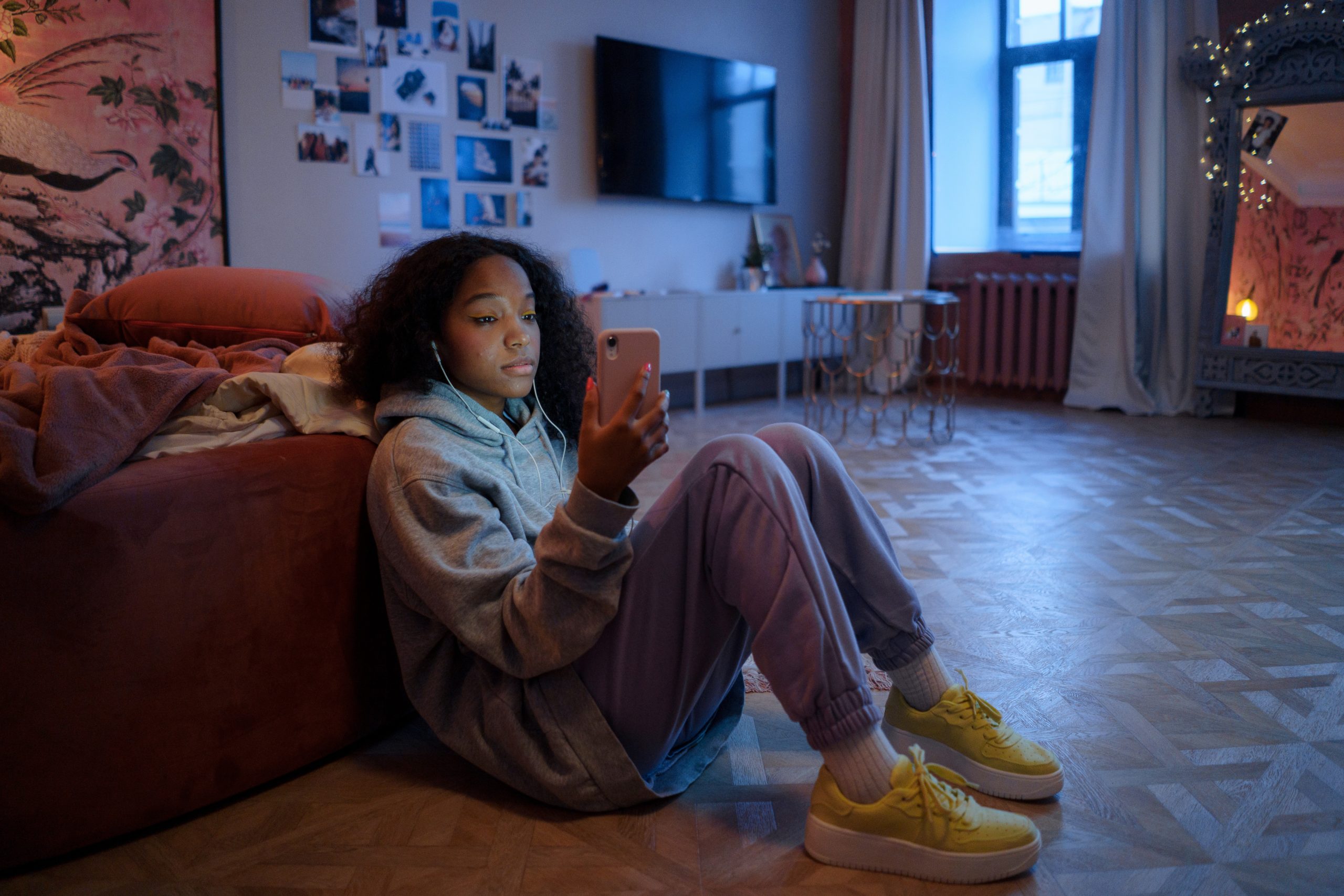 Young Woman Sitting on Floor in Cozy Room Looking at Phone While Wearing Earbuds.