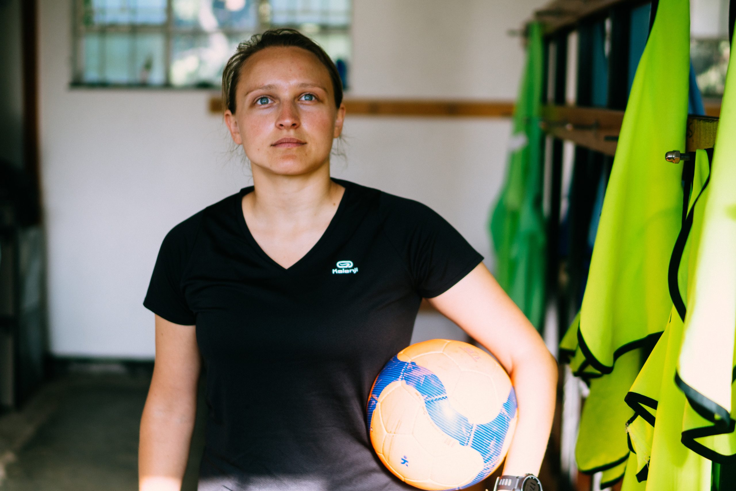 Young woman standing in a locker room holding an orange blue striped soccer ball under her arm
