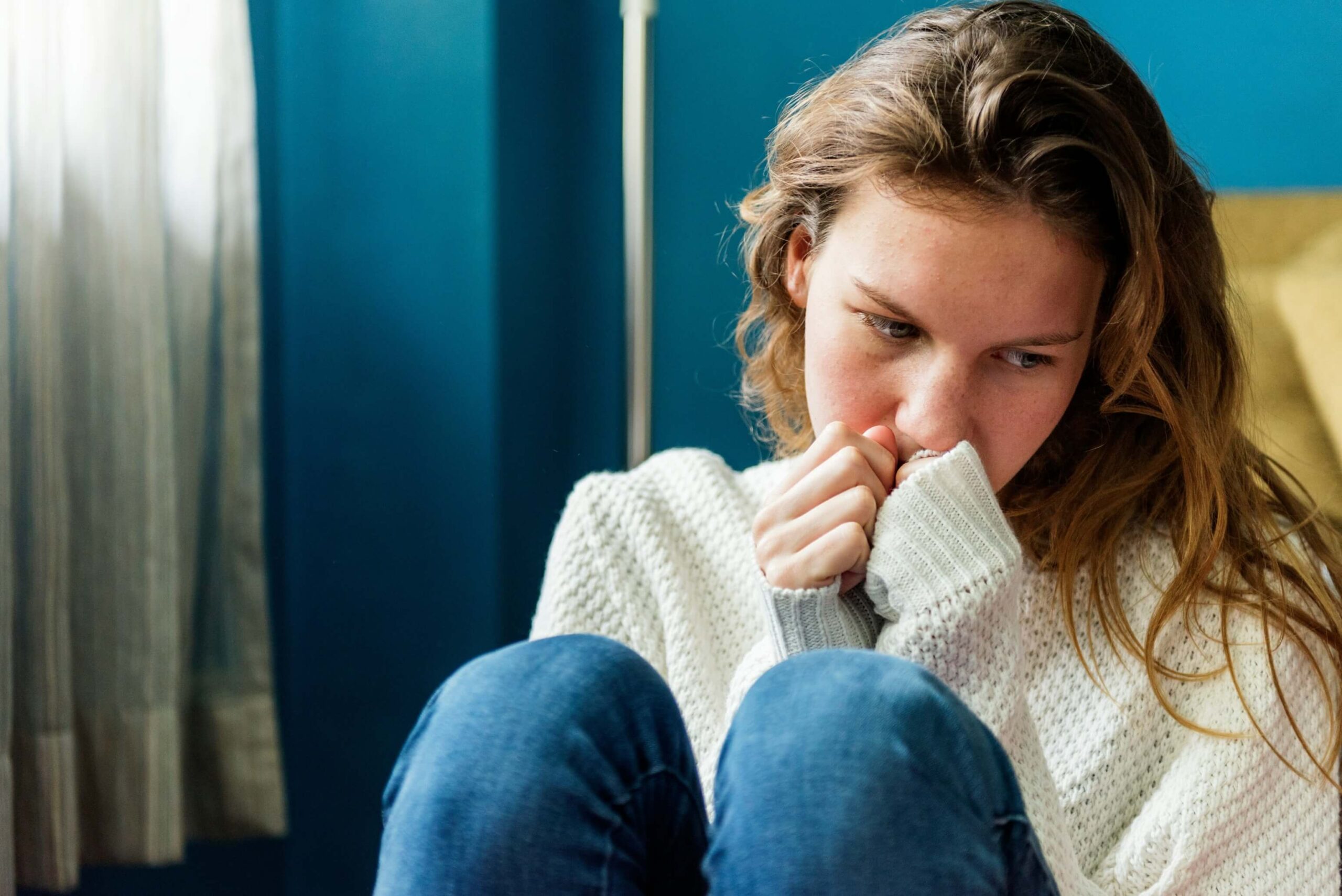 A young person wearing a white sweater holds their hands to their mouth and looks down with a sad expression.