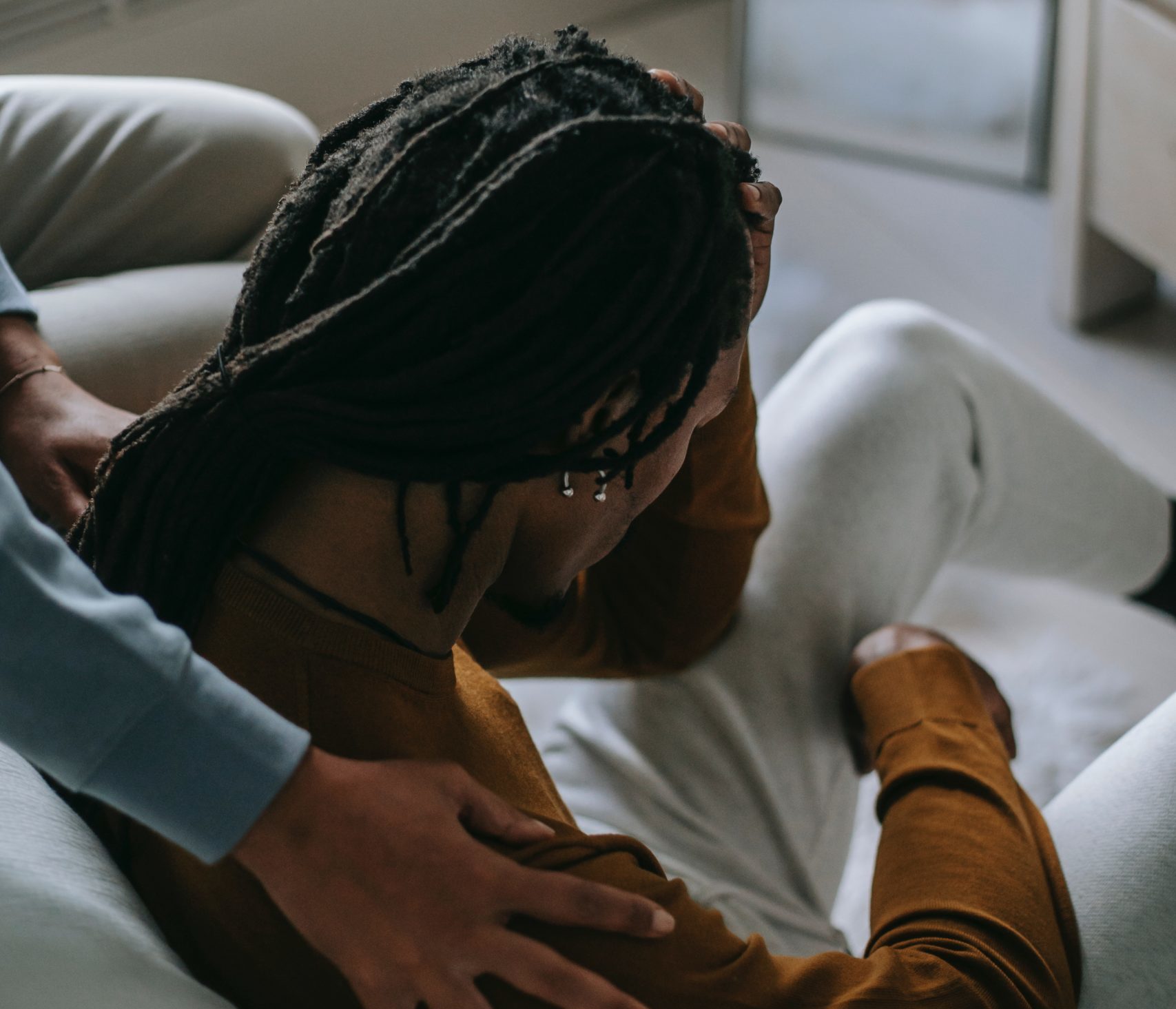 guy with locs sitting on the floor with hand on head and another person behind him comforting him