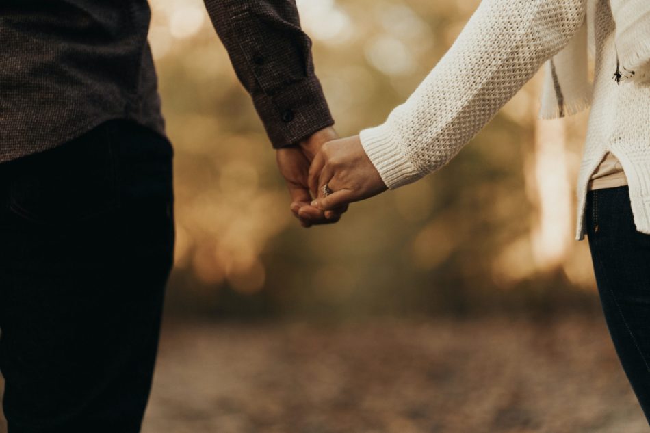two people holding hands, one arm with black shirt, one arm with cream colored shirt, trees and leaves are in the background