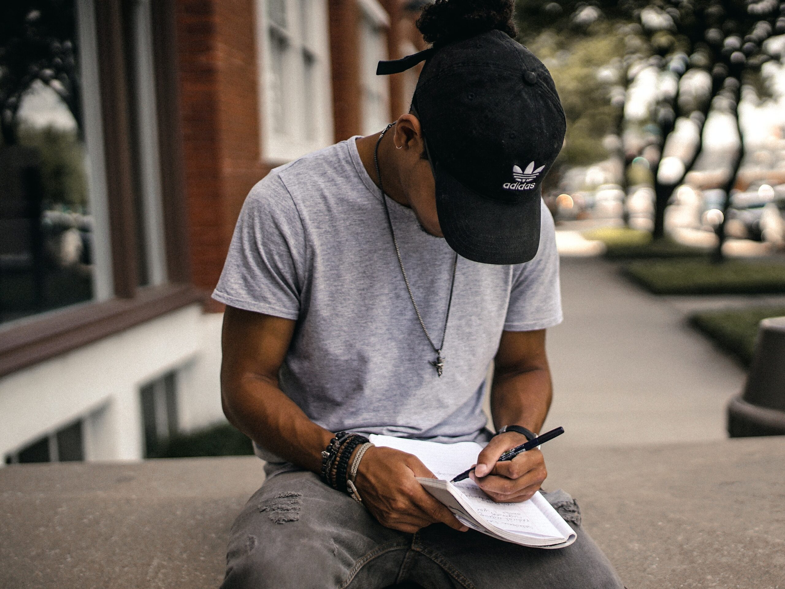 Young Man Wearing Black Cap Sitting Outside Writing Notes in a Notebook.