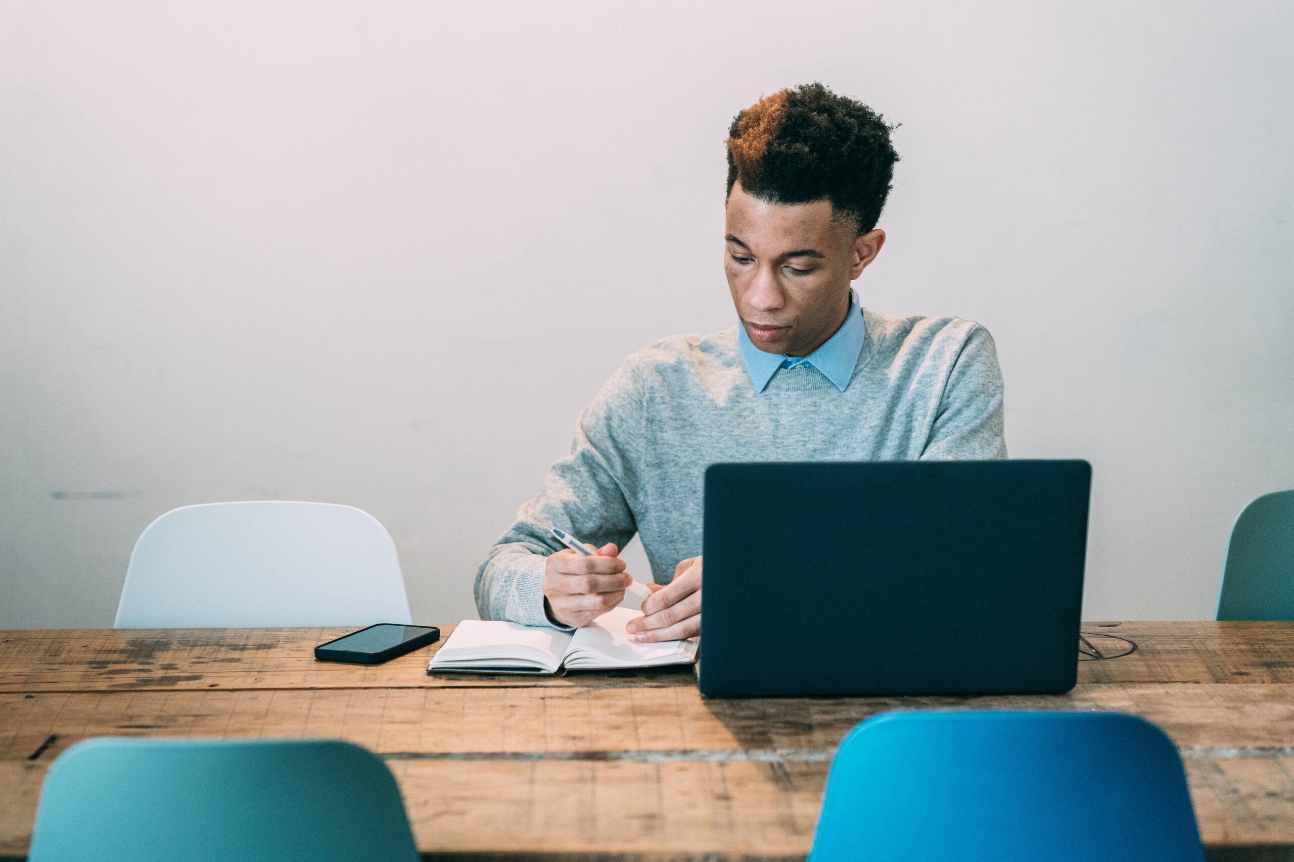 young man sitting at a table with laptop in front of him, writing in a notepad and cell phone on the table besides his notepad
