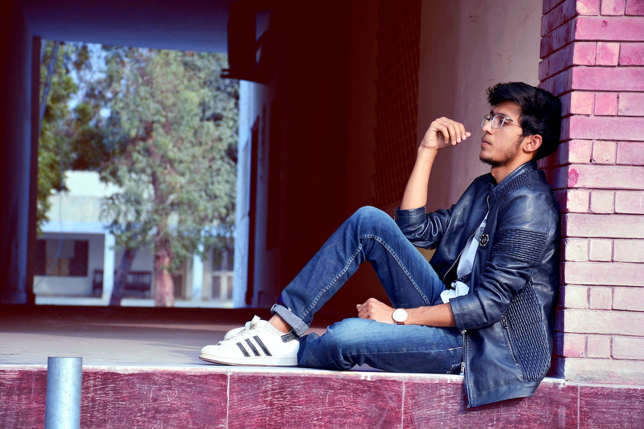 young man sitting outside, leaned up against a wall, deep in thought. residential area and trees in the background