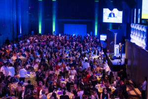 people sitting at tables in ballroom facing the stage where speakers are standing and a person is on a screen