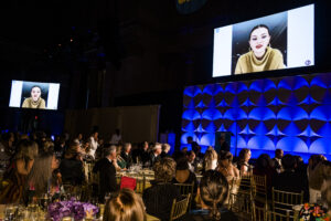 Selena Gomez delivering a message to guess on screen facing an audience of people in ballroom