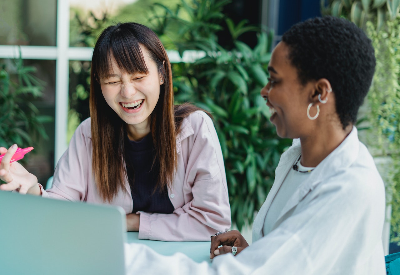 two students of color laughing while sitting at a table with a laptop facing them