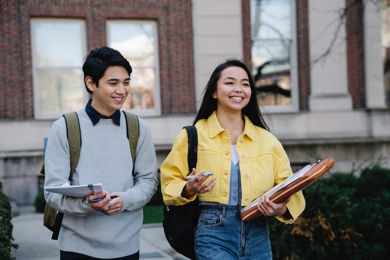 two smiling students connecting outside of the classroom. Students are walking and talking on campus holding their class notes and folders