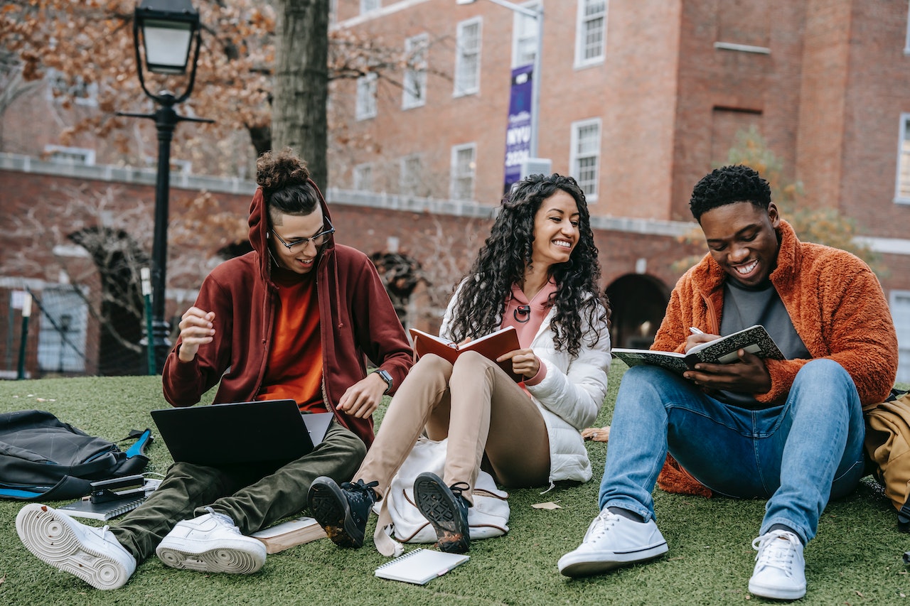 three students sitting on campus lawn talking. reading books and looking at a laptop