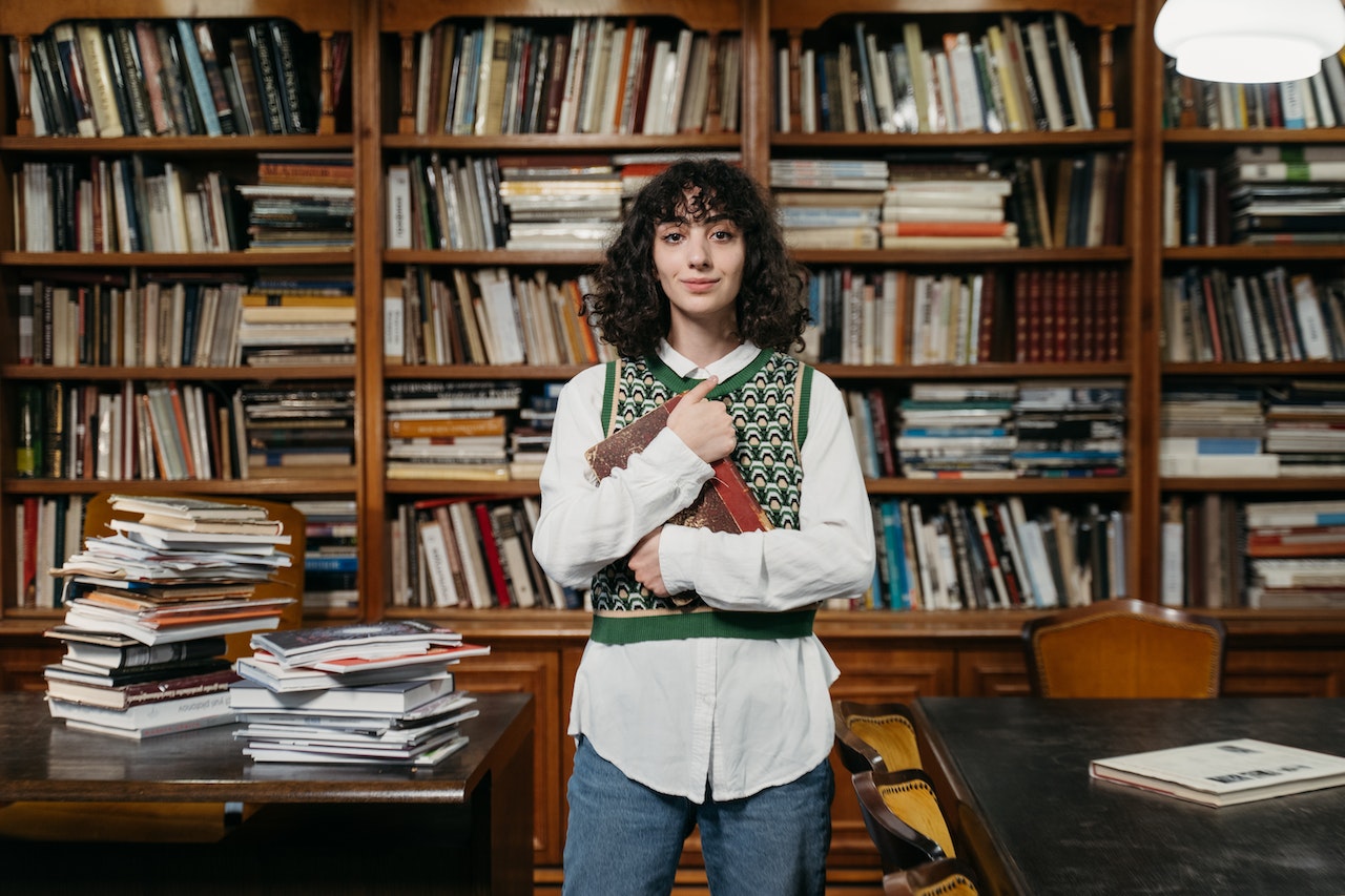 college student standing in library room holding a book with tons of books on tables and shelves behind her