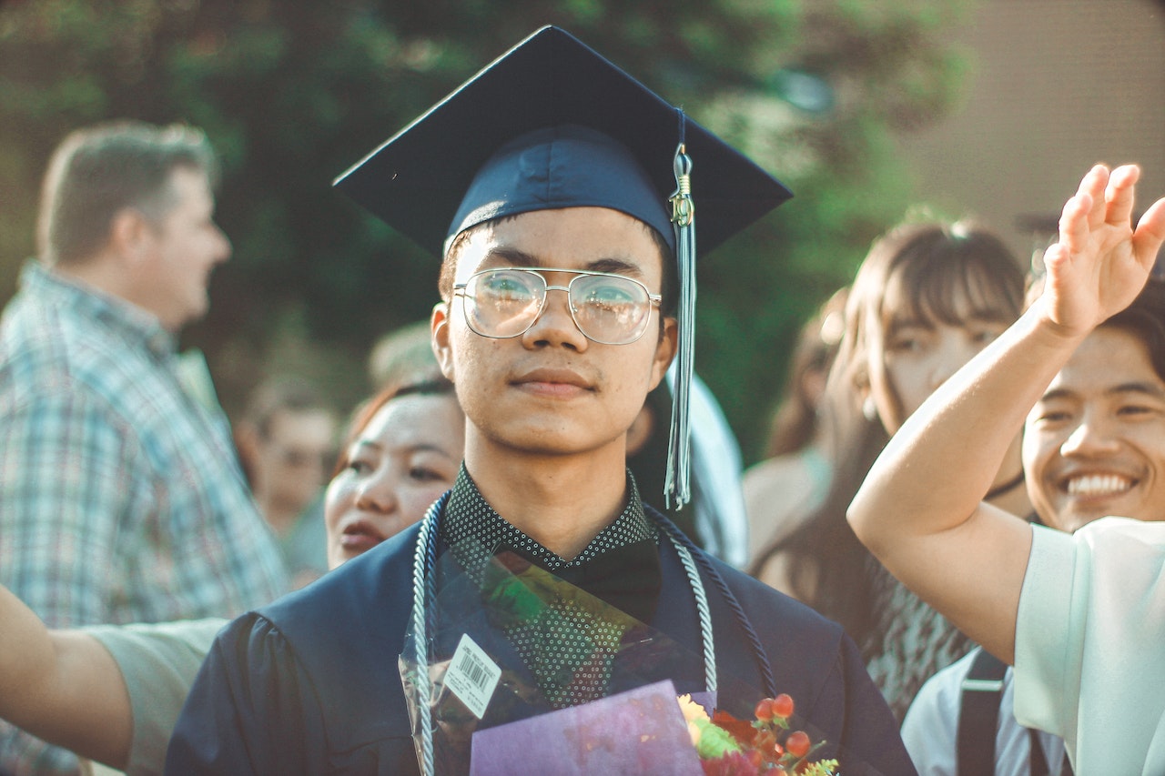high school student at graduation wearing cap and gown and holding flowers with a pensive look on his face