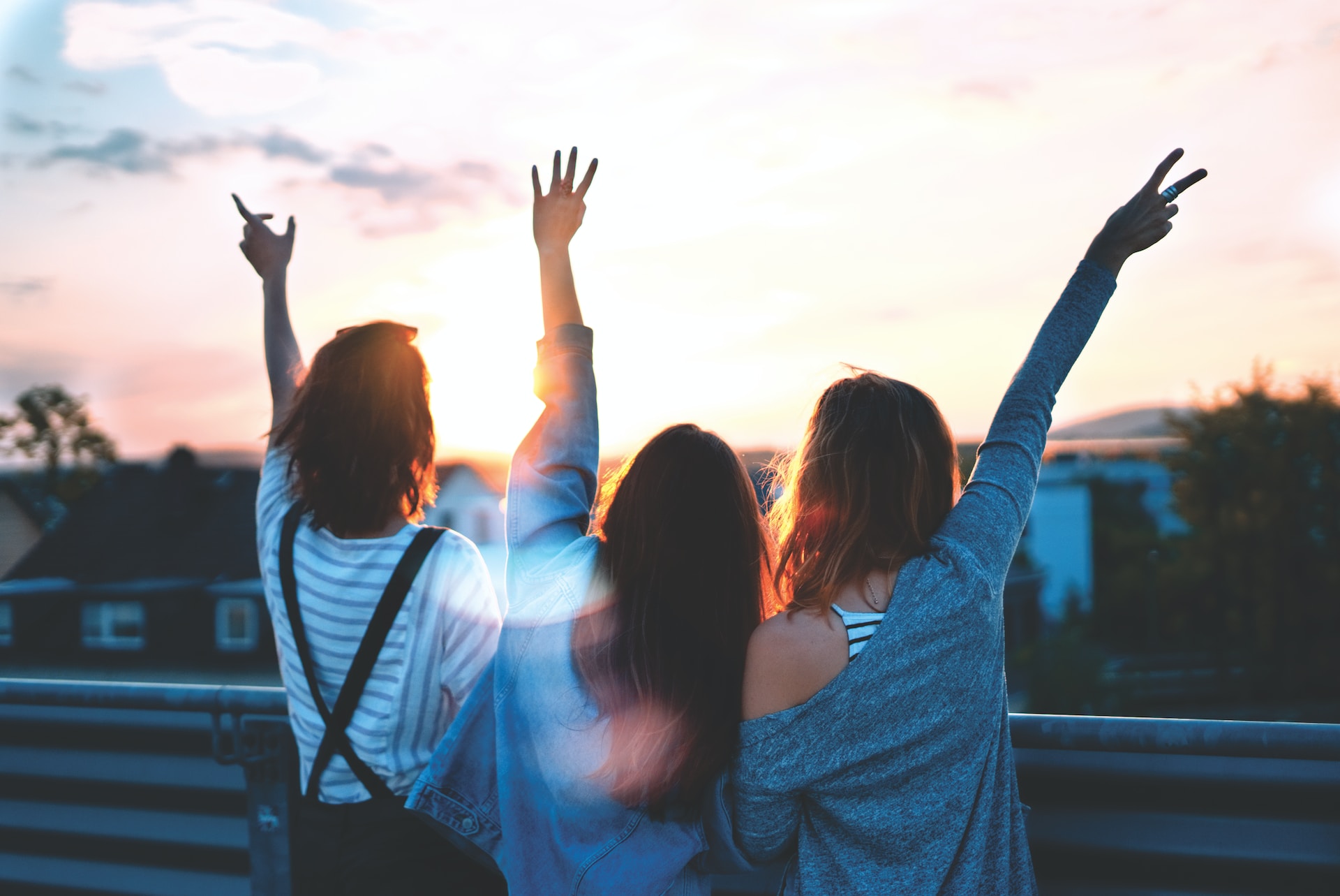college friends standing together cheerfully on a bridge overlooking homes each with one arm held high in the air looking into the sunset