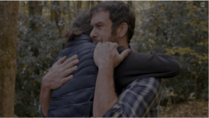 two men hugging with one man looking very upset and trees in the background
