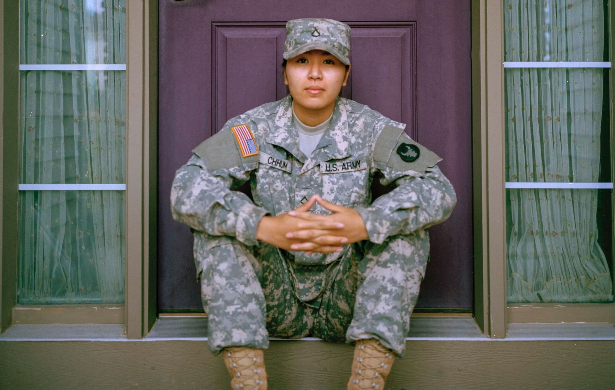 A former US military member sits in front of a door with their arms resting on their knees.