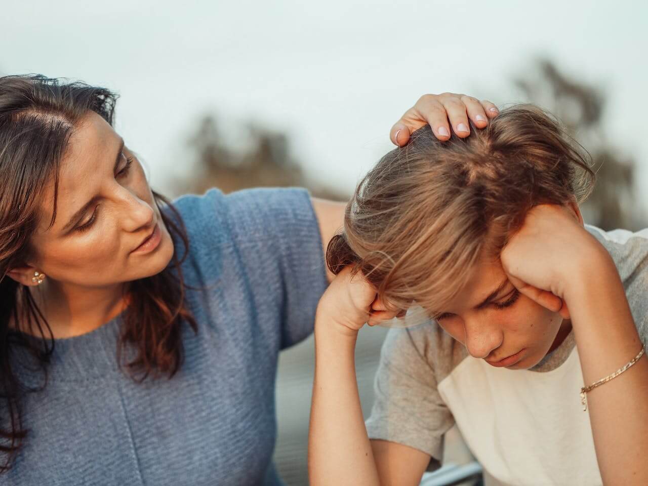 Parent sitting with teen outside patting his head while he leans overs with both fists pressing on his temples
