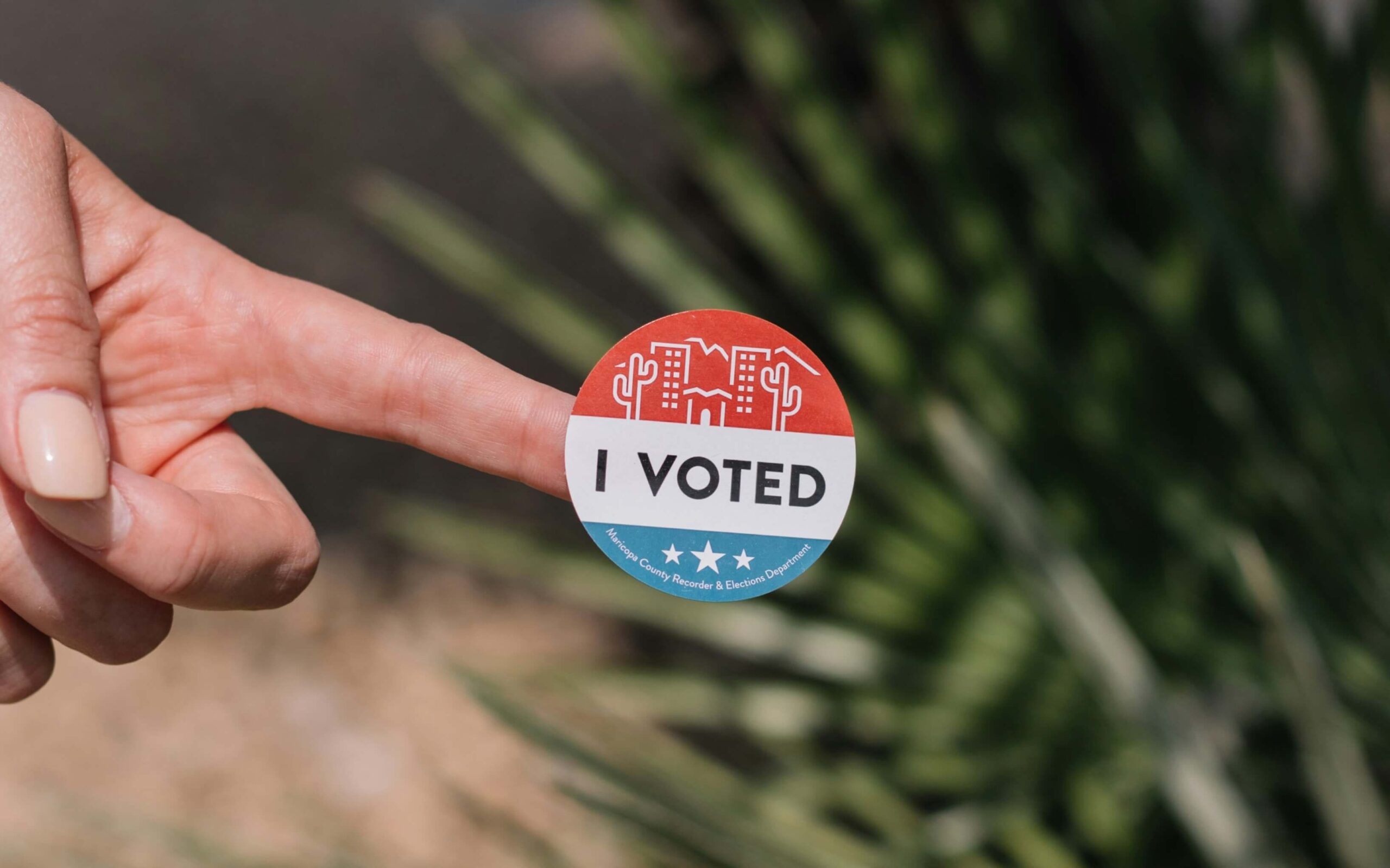 An outstretched hand showing a sticker that reads "I Voted" in red, white, and blue colors.