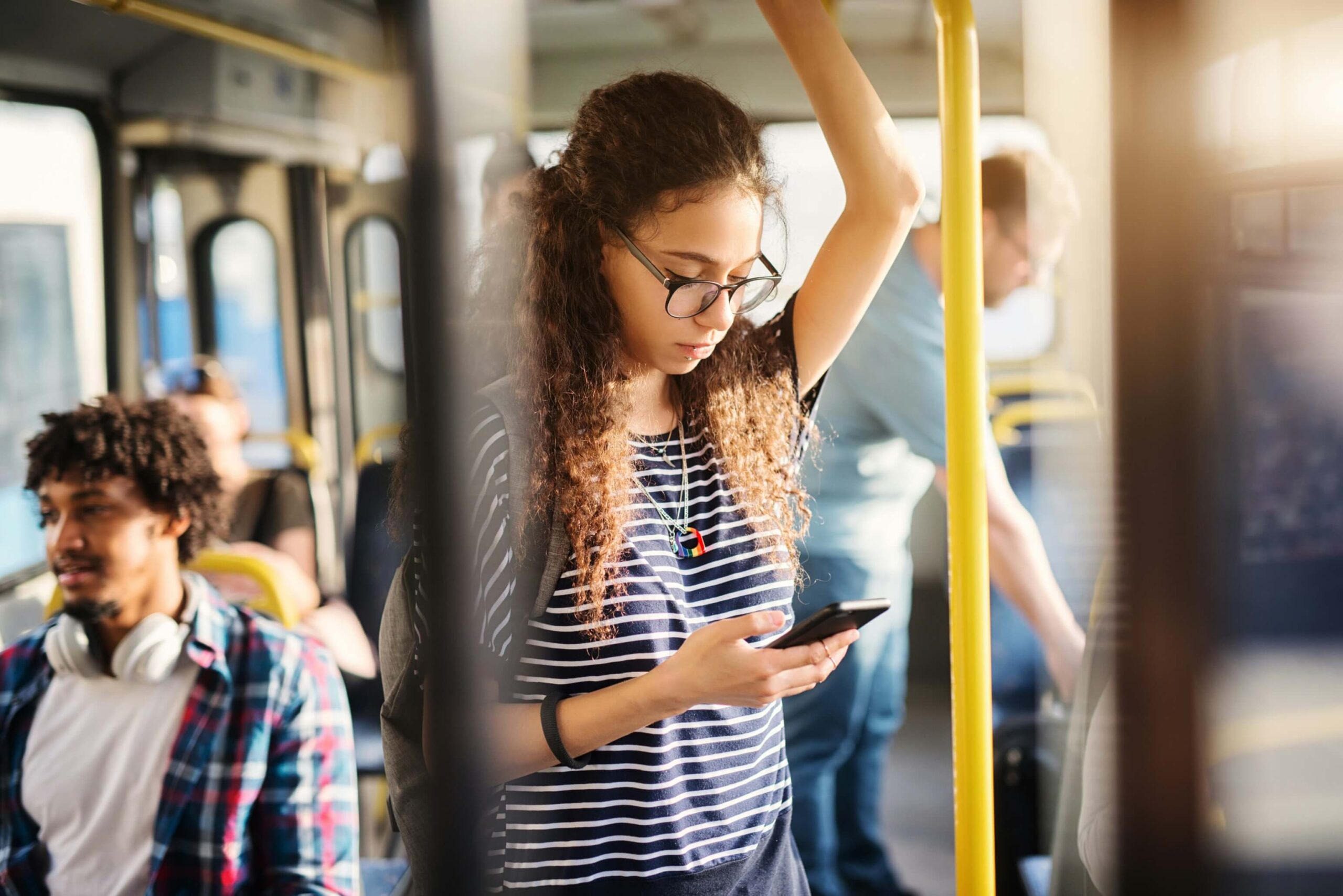 A teen looks down at their phone while holding a handle on a bus.