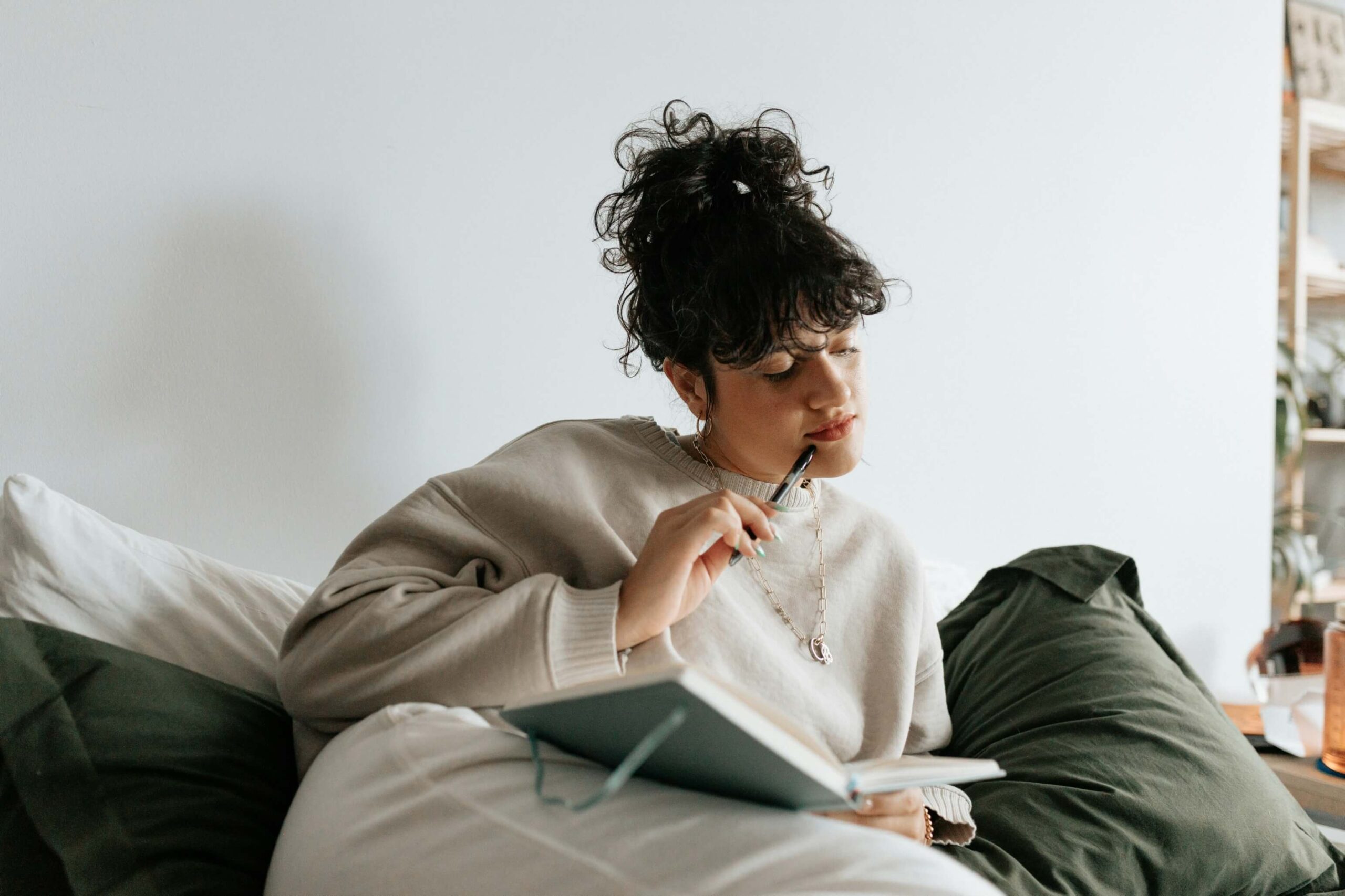A young person sits on a bed with green pillows, holding a pen to their face and looking down at a journal.