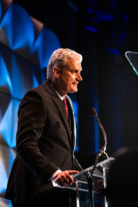 man in black suit standing on stage speaking into a microphone