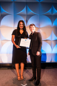 smiling young woman and smiling woman standing side by side on stage with the young woman holding a rectangular shaped award in her hand