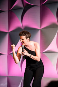 smiling person in black jumper leaning forward while speaking into the microphone in their hand