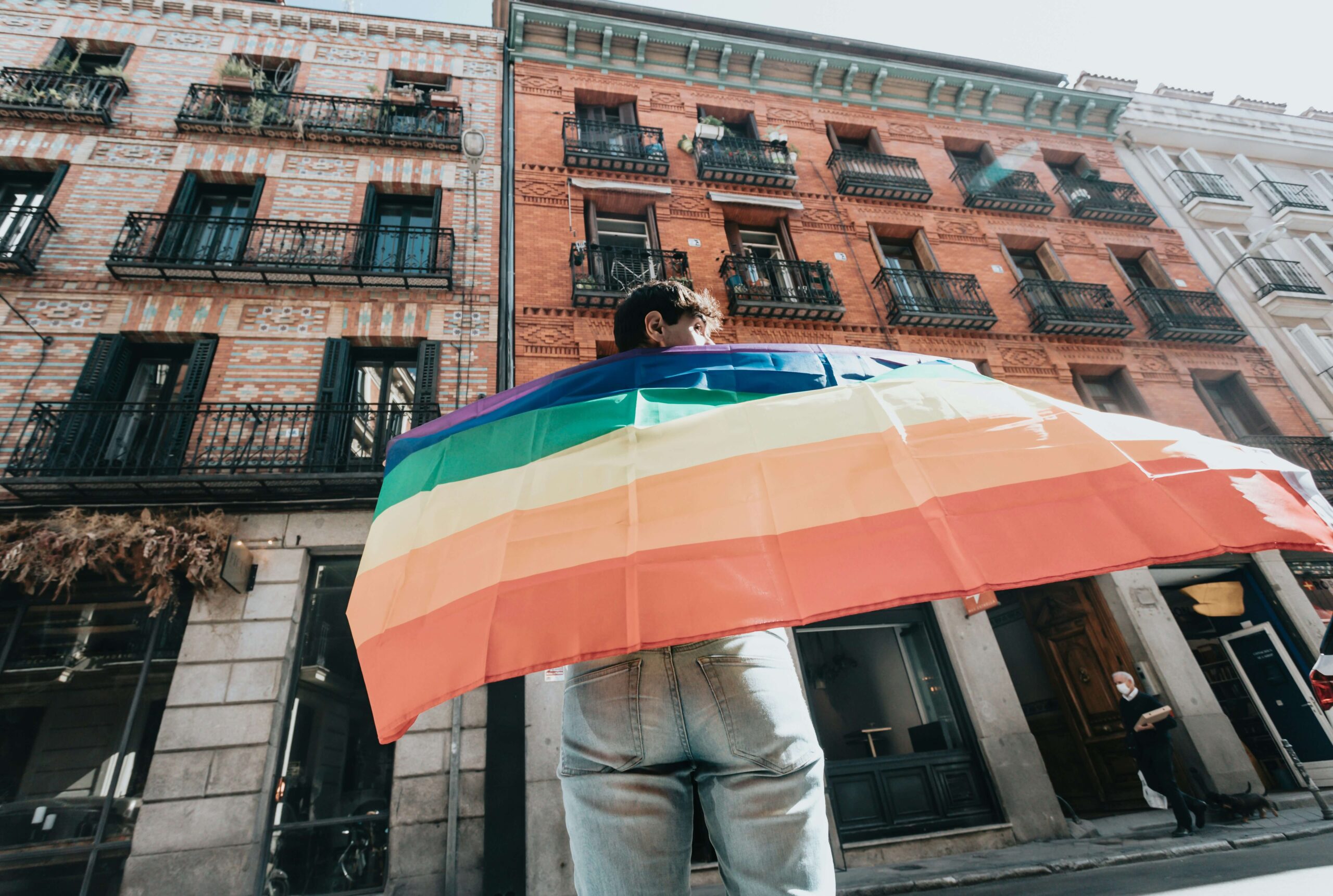 A young person holding a pride flag on their back standing on a city street.