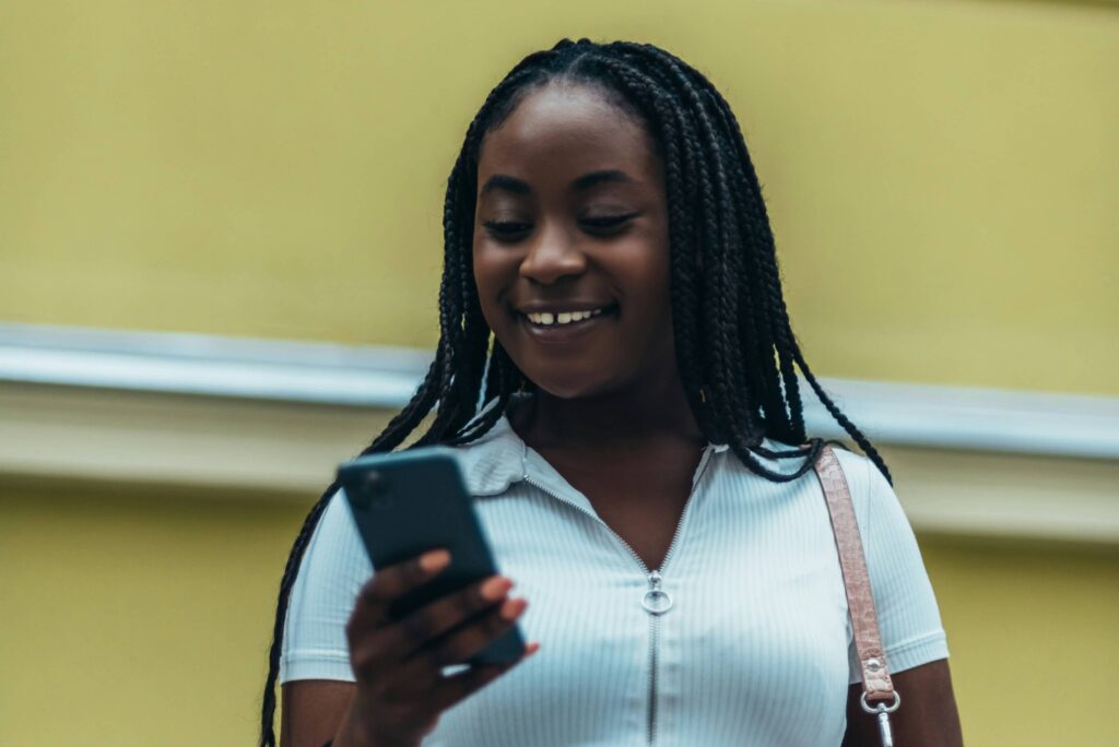 A teen stands outside smiling while looking at their phone in their hand.
