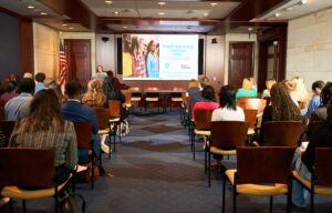 Group of attendees sitting in a room listening to a speaker discuss Youth Advocacy Coalition with Youth Advocacy Coalition presentation on the screen.