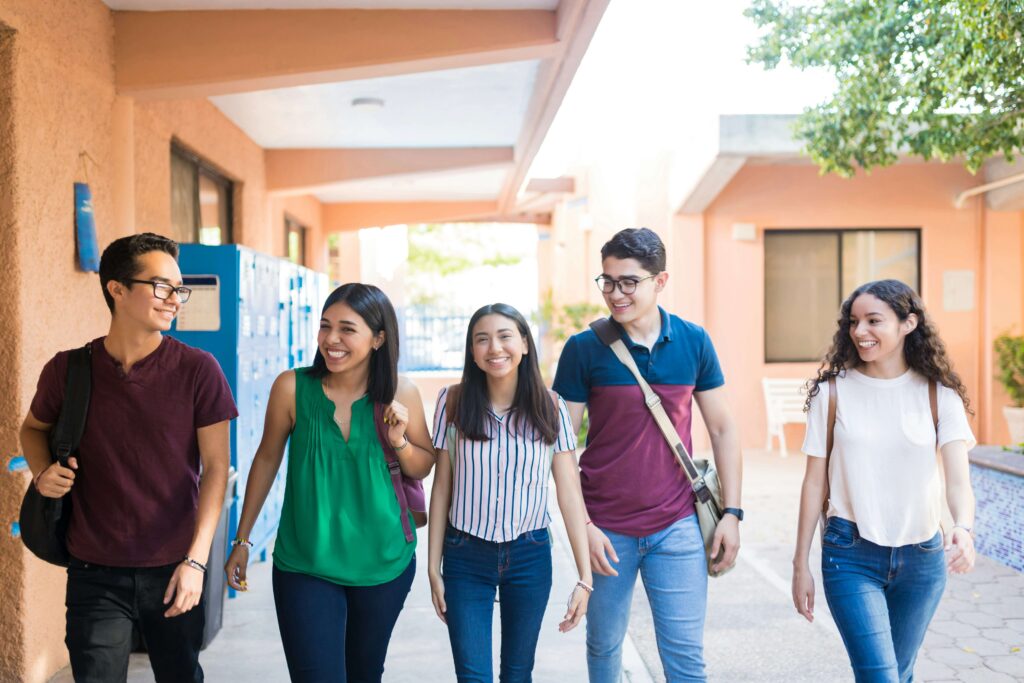 High school students carrying backpacks walk together past a locker bay outside.