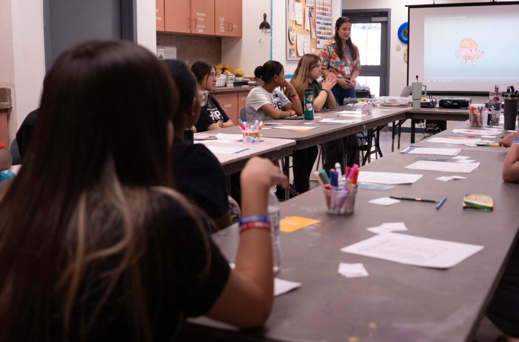 A student sits at a long table covered in papers and markers in a classroom.