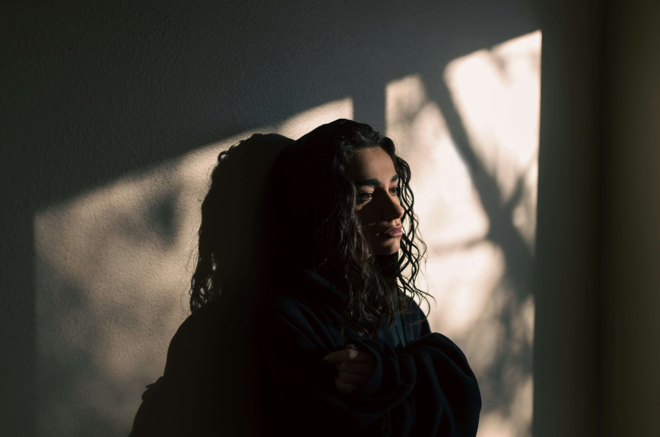 A young person with a sorrowful expression sits in a dark room with light streaming in.