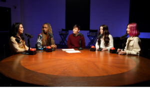 5 people sitting around a table with microphones in front them speaking to one another