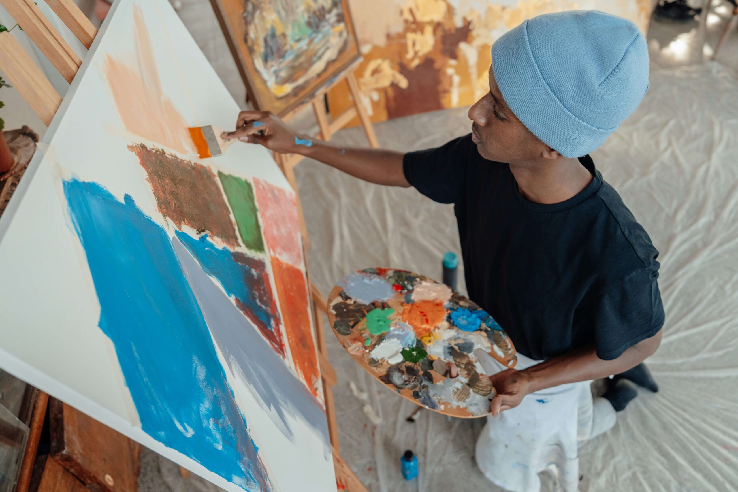 A young person wearing a blue beanie holds an artist's palette while painting an abstract design.