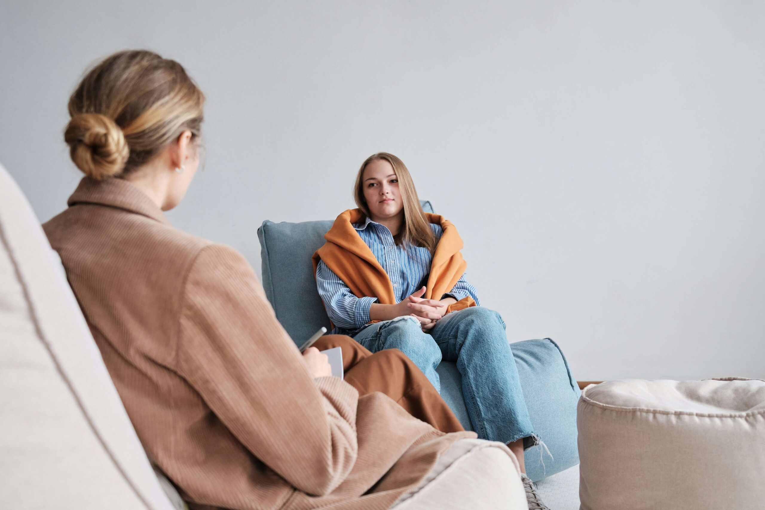 A young person with a blanket wrapped around their shoulders sits and talks to a mental health professional in a cozy room.