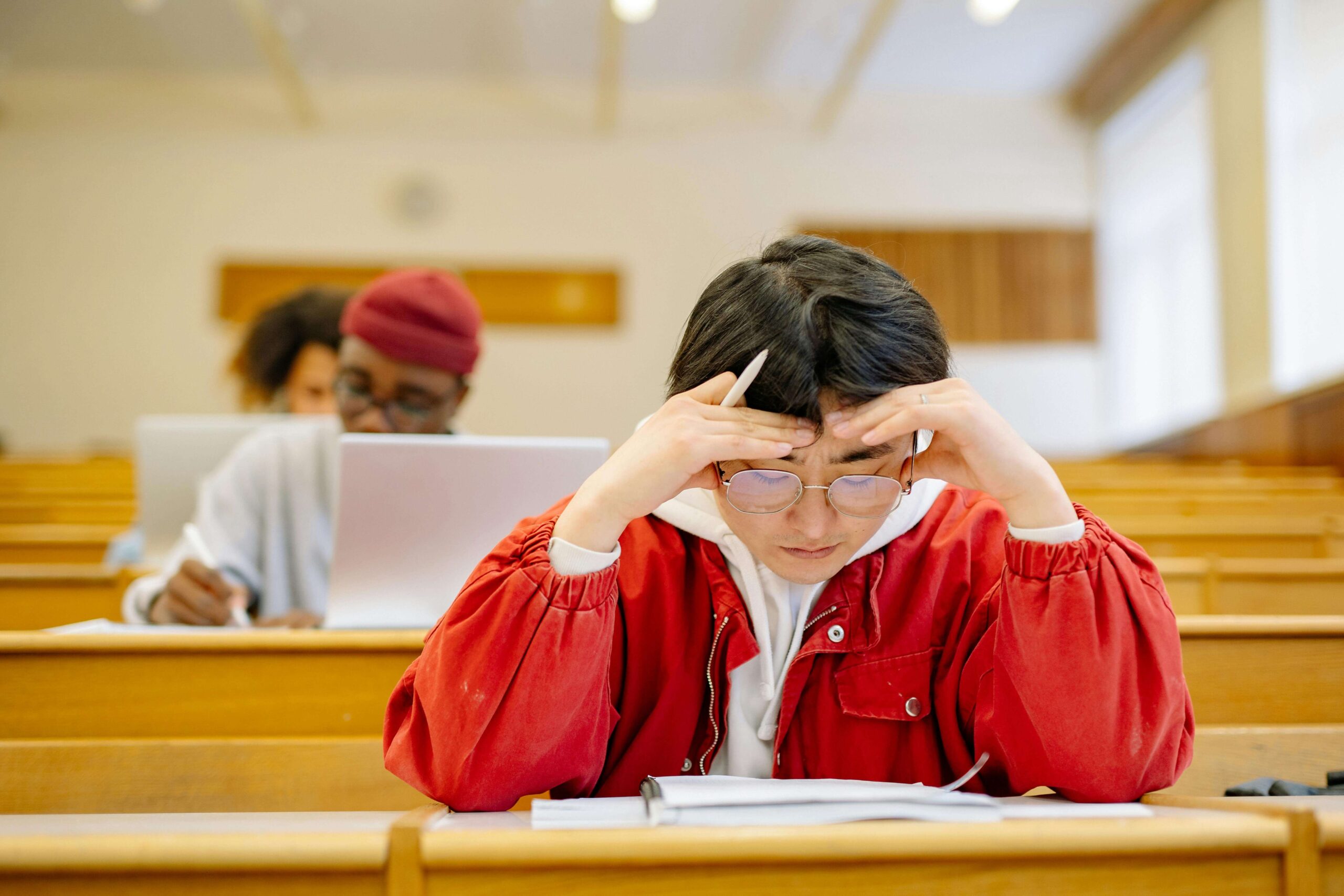 A student sits in a lecture hall, sitting with their head in their hands as they look down at an assignment with a stressed expression.