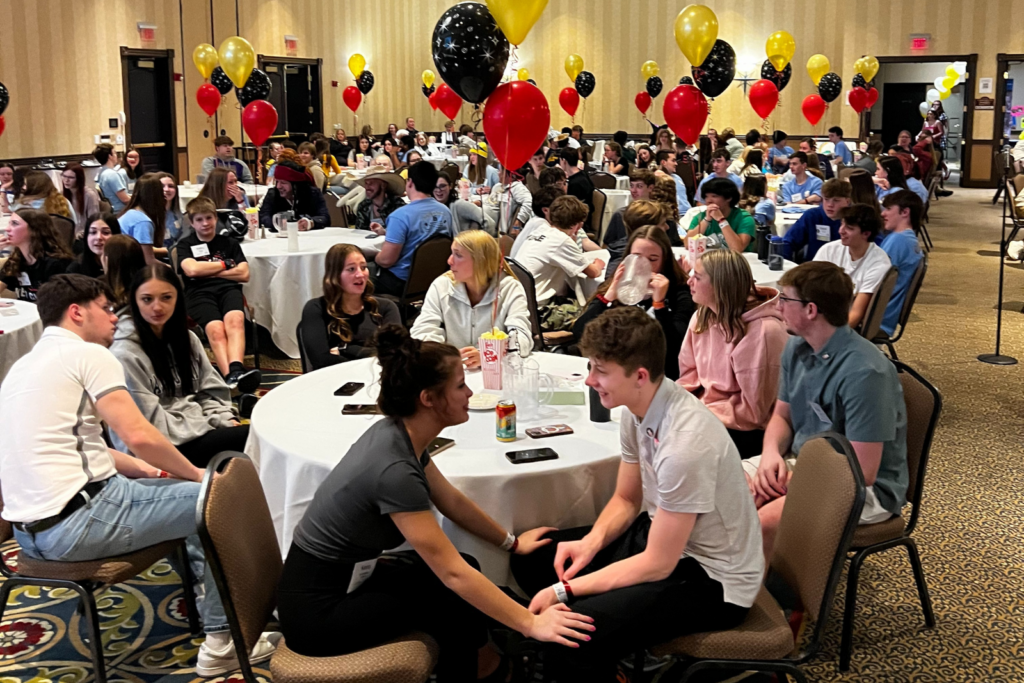Dozens of students are seated and engaged in conversation at round tables during the Wisconsin-Upper Michigan Key Club District Convention (DCON). The packed room includes red, black, and yellow balloons on each table.