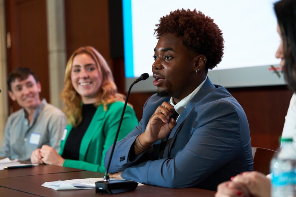 Young man in blue suit sitting at a desk speaking into a microphone while panelists look on.