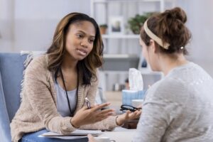 Serious female counselor gestures while talking with Caucasian female client. The counselor is holding eyeglasses and a pen. They are discussing serious issues.